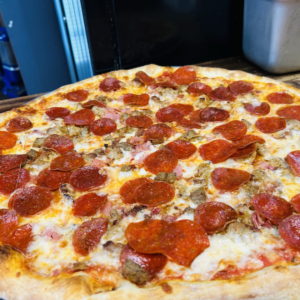 Large thin-crust pizza topped with pepperoni, sausage, ham, mushrooms, and melted cheese on a wooden counter.