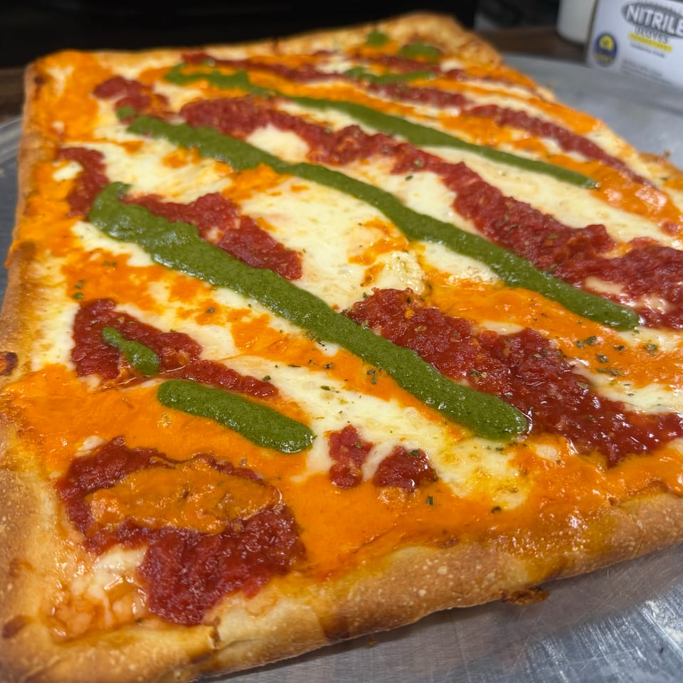 Rectangular pizza with melted cheese, tomato sauce, cured meat, and green pesto drizzled across the surface on a metal tray.