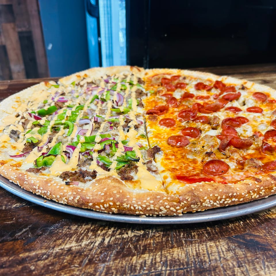 Large sesame-crust pizza split between pepperoni and sausage-vegetable toppings, served on a metal tray atop a wooden table.